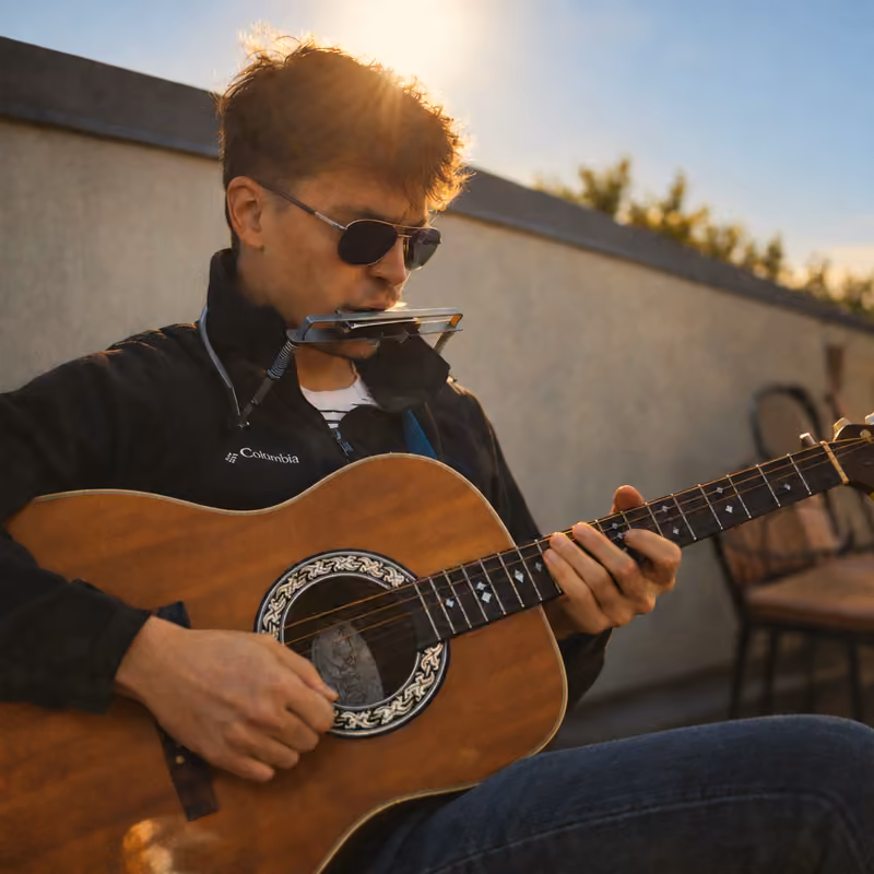 Andrew Wolverton playing acoustic guitar and a harmonica in a neck holder outdoors in warm evening sunlight